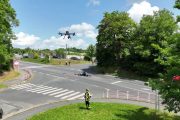 Pilot dronu stojící na travnatém plácku vedle křižovatky. Drony se vznáší nad ním.