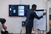 Muž v obleku na poradě stojí před tabulí a vysvětluje kolegům grafy