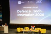 Připrava konference Defence Tech Innovation 2026. Žlutá křesla a stolečky před velkým plátnem. Bokem je umístěný řečnický pult.